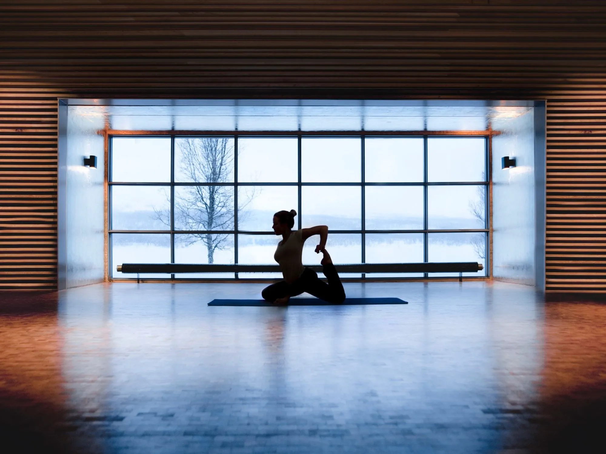 Person som utför yoga framför ett stort fönster med utsikt över ett snötäckt landskap.