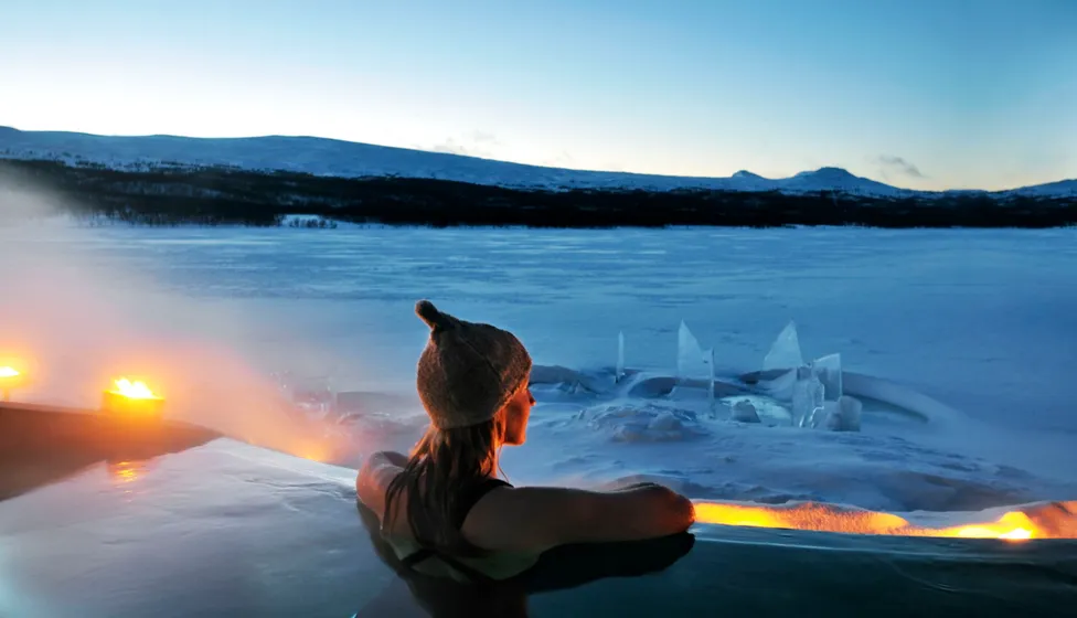 Person som sitter i varmt bad utomhus och tittar ut över isbelagd sjö och fjäll.