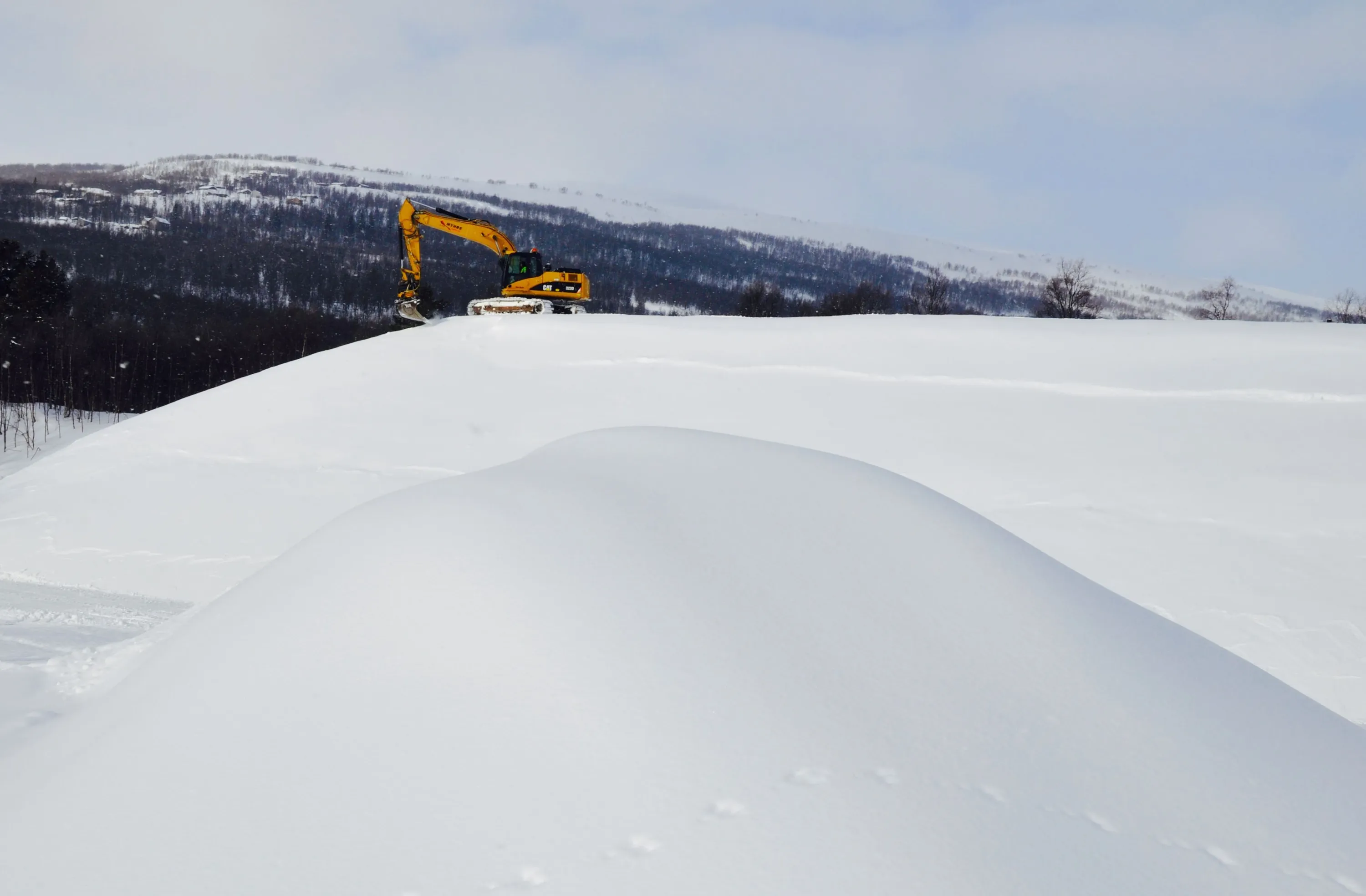 Grävmaskin som gräver i snön, fjäll i bakgrunden.