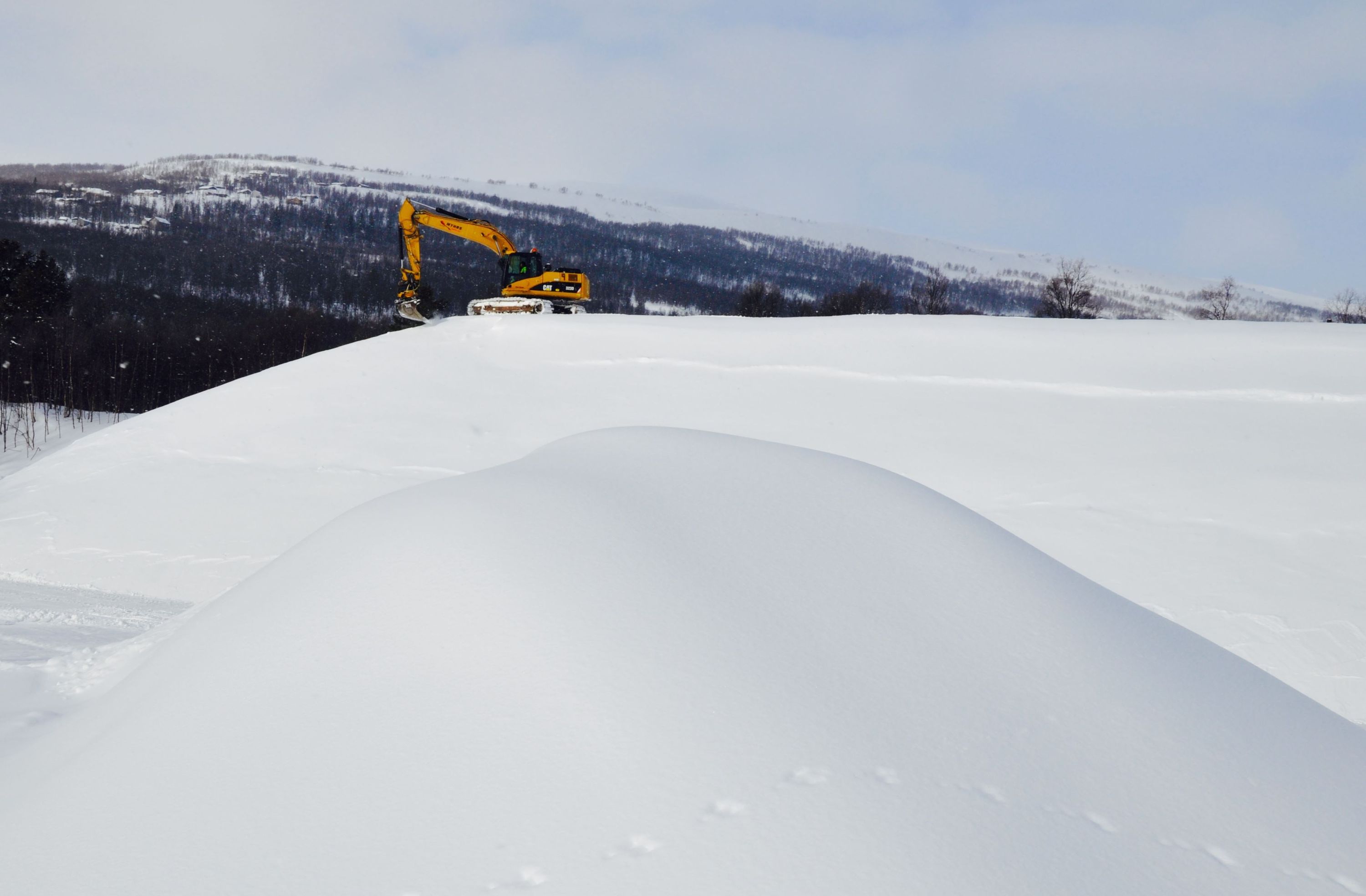Grävmaskin som gräver i snön, fjäll i bakgrunden.
